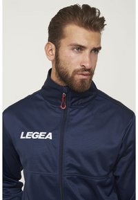 Chaqueta de pista azul marino hecha de tejido suave, con cuello alto, cierre de cremallera con tirador rojo y logo blanco "LEGEA" en el pecho.