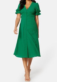 Vestido midi verde con mangas cortas abullonadas, escote en V y detalles de botones en la parte delantera. Tejido suave con una ligera textura.