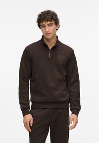 Hnedý zip-up sveter vyrobený z textúrovanej tkaniny má malú nášivku na hrudi a rebrované manžety. Kombinovaný s ladiacimi nohavicami.