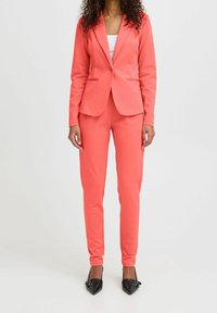 Costume deux pièces corail avec un blazer ajusté et un pantalon droit. Le tissu est lisse, avec des lignes épurées et un minimum de détails. Associé à des escarpins noirs pointus.