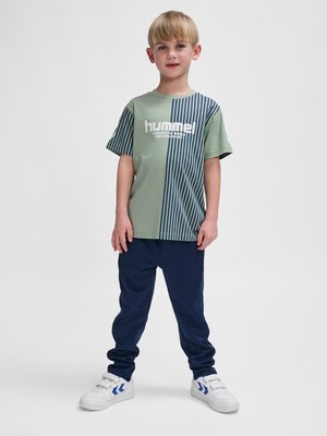 Ung dreng stående, iført grøn og stribet hummel t-shirt, navy bukser og hvide sneakers med blå Chevron-logoer, ensfarvet lys baggrund.