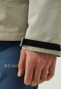 Bež vodootporna jakna sa rukavima, koji imaju debele crne čičak manžete, nosi se preko plave tkanine s logotipom. Ruka se blago oslanja na tkaninu.