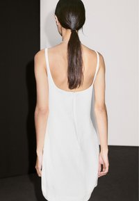 Weißes ärmelloses Kleid mit tiefem Rückenausschnitt, das ein einfaches Design und eine glatte Textur aufweist. Der Stoff wirkt leicht und fließend.