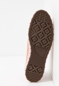 Semelle en caoutchouc marron d'une chaussure Converse All Star avec des motifs géométriques de crampons et le numéro de pointure 5 visible sur une tige en tissu rose clair.