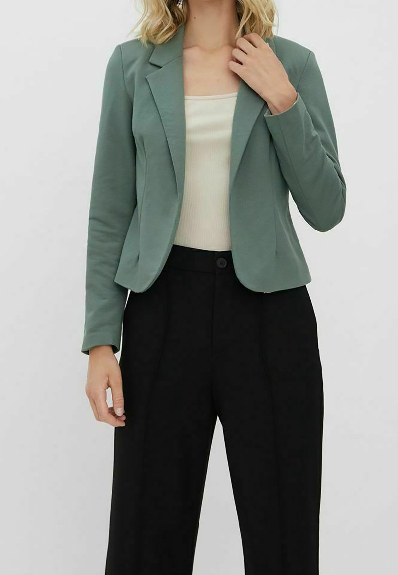 Femme portant un blazer vert sauge, un débardeur côtelé crème et un pantalon large noir taille haute sur fond blanc.