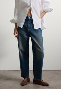 Jeans de mezclilla azul claro con cintura alta, pierna recta y un ligero desgaste. Combinados con una camisa blanca de botones de corte holgado.