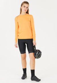 Långärmad orange träningsskjorta i kombination med svarta cykelshorts. Buren med svarta strumpor och skor, håller en svart hjälm.