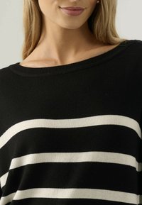 Primo piano di una persona che indossa un maglione nero con ampie righe orizzontali bianche e un'ampia scollatura, che mostra le clavicole e le labbra.