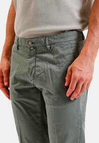 Pantaloni grigio-verde chiaro realizzati in un tessuto liscio, con chiusura a bottone, tasche laterali e una vestibilità rilassata. Dettaglio della trama evidente.