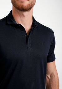 Mornarsko polo majica z teksturiranim pletenjem, z dvema gumboma na zapenjanju in vezenim logotipom na levi strani prsnega koša. Kratke rokave z zloženimi manšeti.