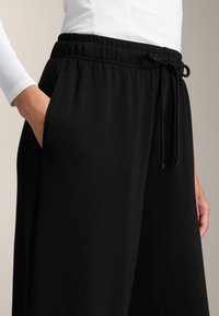 Zwarte wijde broek met een elastische tailleband en een verstelbaar trekkoord. Bevat zijzakken en heeft een soepele, zachte stoftextuur.