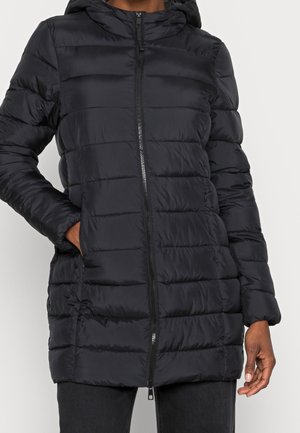 Doudoune matelassée noire avec capuche, fermeture éclair frontale et poches latérales portée sur un pantalon sombre, main du modèle dans la poche, sur fond blanc.