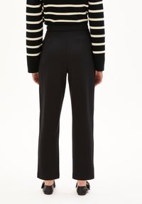 Personne portant un pantalon noir à jambes droites, des chaussures noires et un haut rayé noir et blanc à manches longues, se tenant dos à un fond blanc.