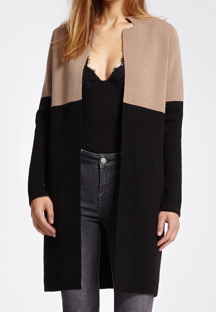 Femme portant un long cardigan color-block beige et noir par-dessus un top noir avec bordure en dentelle et un jean gris foncé, debout devant un fond blanc.