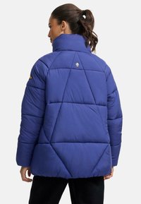Lilla puffer jakk, millel on pikk krae, tikanditunne ja logode detail taga. Sile tekstuur ja lõdva istuvusega. Mustad aktsendid.