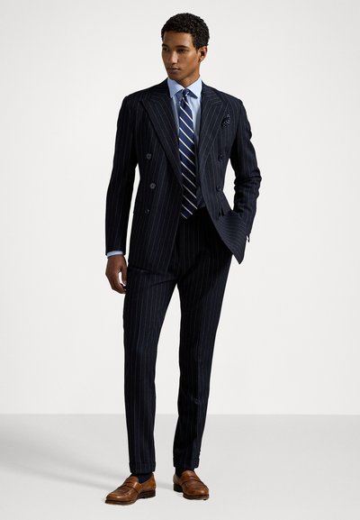 Fato duplo abotoado com riscas em azul-marinho, camisa azul clara e gravata riscada. Sapatos de pele castanha completam o visual formal.