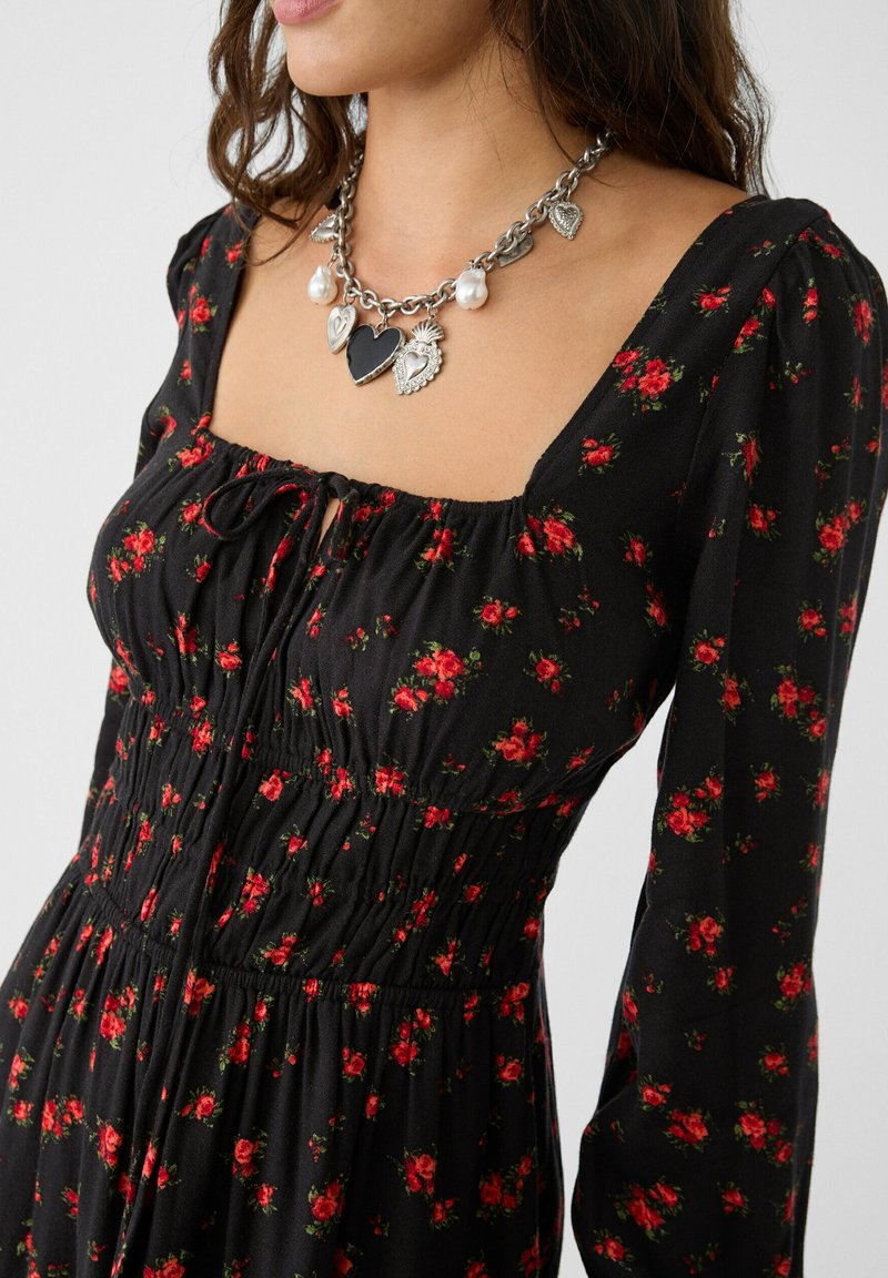 Schwarzes Kleid mit rotem Blumenmuster, gerafftem Oberteil, langen Ärmeln und quadratischem Ausschnitt. Mit einer geschichteten metallischen Herz-Halskette mit Perlen.
