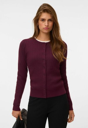 Femme aux longs cheveux bruns portant un cardigan bordeaux en maille torsadée boutonné et un pantalon noir, tenant une veste noire dans sa main gauche.
