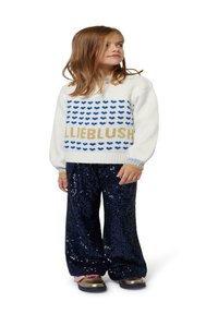 Witte sweater met blauw hartpatroon en "LIEBLUSH" in goud, gecombineerd met marineblauwe pailletten wijde broek en schoenen met gouden accenten.