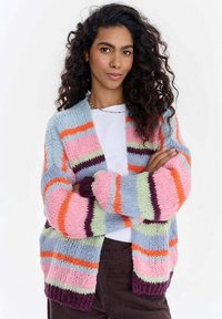 Žena s dlouhými vlnitými vlasy má na sobě vícero barevné pruhované pletené sako přes bílé tričko a stojí s překříženýma rukama proti jednoduchému pozadí.