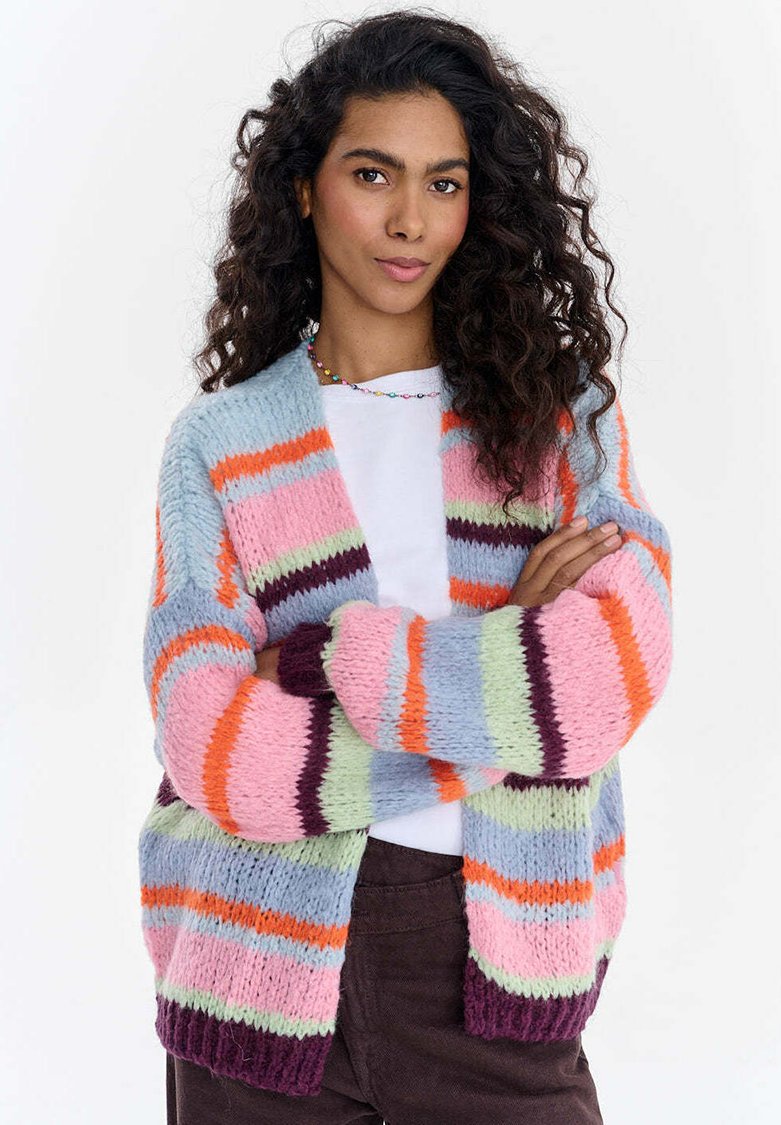 Žena s dlouhými vlnitými vlasy má na sobě vícero barevné pruhované pletené sako přes bílé tričko a stojí s překříženýma rukama proti jednoduchému pozadí.