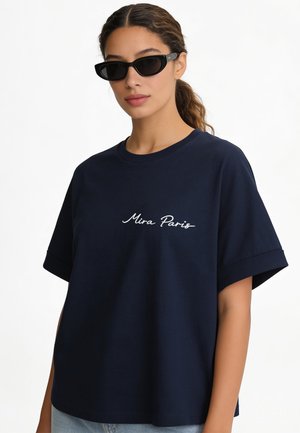 Vrouw met een zwarte zonnebril en een donkerblauw T-shirt met "Mira Paris" geschreven in witte cursieve tekst.