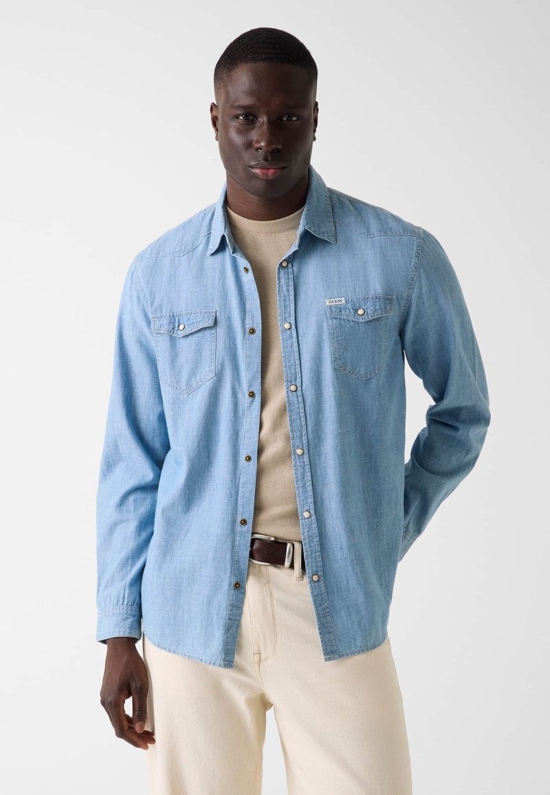 Homme debout devant un fond uni, portant une chemise en jean bleu clair sur un pull beige, un pantalon crème et une ceinture marron foncé.