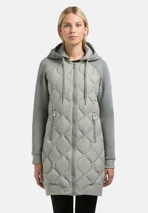 Kobieta ubrana w długą, jasnoszarą pikowaną bluzę z kapturem na zamek błyskawiczny, ze sznurkami i zamykanymi kieszeniami po bokach, na jednolitym tle.