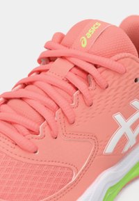 Korallna atletska cipela s mrežastim i sintetičkim gornjim dijelom, bijelim detaljima, ravnim vezicama i zelenim detaljem na potplatu, ističući teksturirane površine i brendiranje.