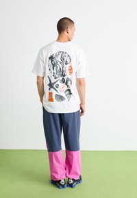 Homme portant un t-shirt blanc avec des motifs abstraits de nature noirs et oranges dans le dos, associé à un pantalon bicolore bleu et rose et des baskets noires.