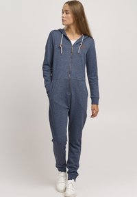 Marineblå jumpsuit laget av myk, teksturert stoff. Har glidelås foran, hette med snor, og sidelommer. Buksene smalner inn ved anklene.