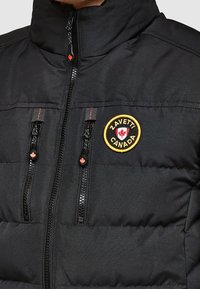 Czarna ocieplana kurtka z wysokim kołnierzem, dwoma przednimi kieszeniami na zamek oraz okrągłym logo z napisem „Zavetti Canada” i liściem klonu.