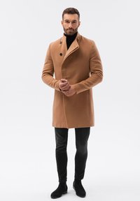 Cappotto di lana color cammello con design doppiopetto, caratterizzato da bottoni neri, indossato sopra un dolcevita nero e abbinato a pantaloni neri e stivali.
