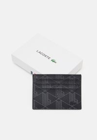 Sort tekstureret kortholder med geometrisk mønster og prægede logo, vist på en hvid æske med Lacoste-brandnavnet.