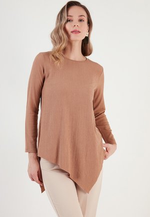 Long-sleeve, textured top in a light brown color with an asymmetrical hem and a round neckline, paired with beige pants.