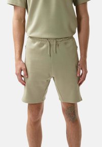 Pantaloni corti verdi di media lunghezza realizzati in tessuto morbido. Presentano un elastico in vita con cordino e tasche laterali. Design semplice, senza motivi.
