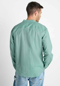 Man in een groen overhemd met lange mouwen en lichtblauwe jeans, met de rug naar de camera met zijn handen in zijn zakken, tegen een effen witte achtergrond.