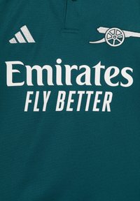 Vihreä polo-paita, joka on valmistettu tekstuurista kankaasta ja jossa on valkoinen "Emirates FLY BETTER" -logo ja hopeiset Adidas-raidat. Mukana on pieni tykki-graafinen kuvasto.