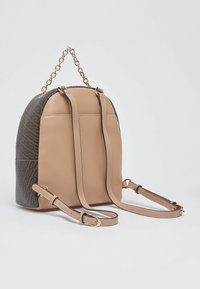 Sac à dos en similicuir beige avec un panneau texturé marron, chaîne dorée et bretelles réglables, forme courbée et couture nette.