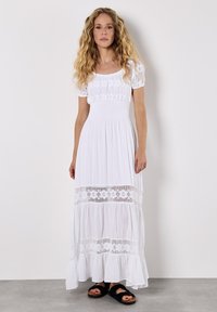 Witte maxi-jurk met puff sleeves, gerimpelde buste, gelaagde gerimpelde zoom en bloemenkantaccenten. Draag met zwarte slide-sandalen.