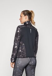 Crni i mramorni sportski jakna s visokim ovratnikom i reflektirajućim detaljem na leđima. Tkanina koja odvodi vlagu, osmišljena za aktivno nošenje.