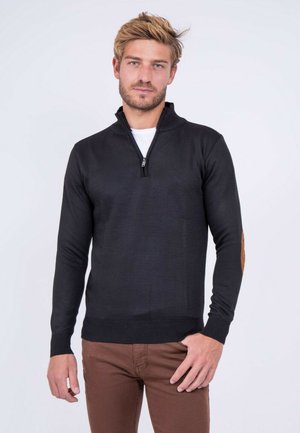 Homme blond avec une légère barbe, portant un pull noir à fermeture éclair partielle avec des coudières et un pantalon marron, debout devant un fond clair uni.