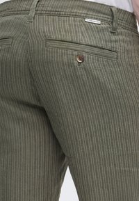 Primer plano de pantalones a rayas verdes con un bolsillo trasero abotonado y una pequeña etiqueta blanca de la marca encima del bolsillo.