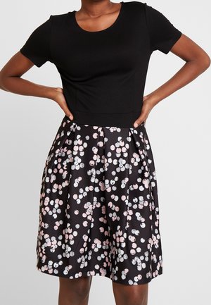 Vrouw draagt een zwart topje met korte mouwen en een zwarte rok met een patroon van roze en witte stippen, handen rustend op de heupen.