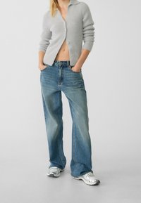 Donna che indossa un cardigan grigio chiaro peloso, jeans blu larghi e sneakers argento, in piedi con le mani nelle tasche contro uno sfondo semplice.
