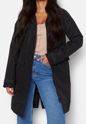 Manteau imperméable noir avec des manches longues, des boutons sur le devant, des poches latérales et une coupe droite. Porté sur un haut léger et un jean bleu.