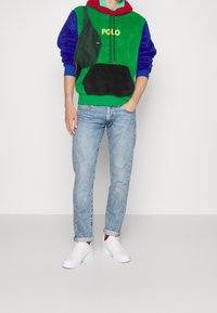 Zelená a modrá fleecová mikina s kapucí s logem "POLO", černá kapsa, doplněná světle modrými džíny a bílými teniskami s červenými akcenty.