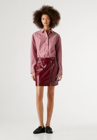 Chemise à boutons rayée rouge avec manches longues, associée à une mini-jupe en cuir bordeaux avec un zip et une finition brillante. Mocassins noirs.