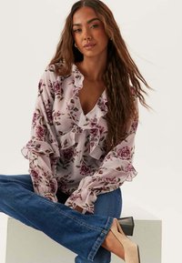 Blommig blus i ljusrosa med volangkrage och ärmar, med mörkrosamönster. Ett par blåjeans och beige högklackade skor.
