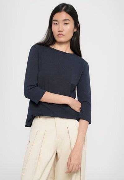 Top blu navy a maniche 3/4 realizzato in tessuto morbido, con scollatura rotonda e taglio rilassato, abbinato a pantaloni beige chiaro plissettati.